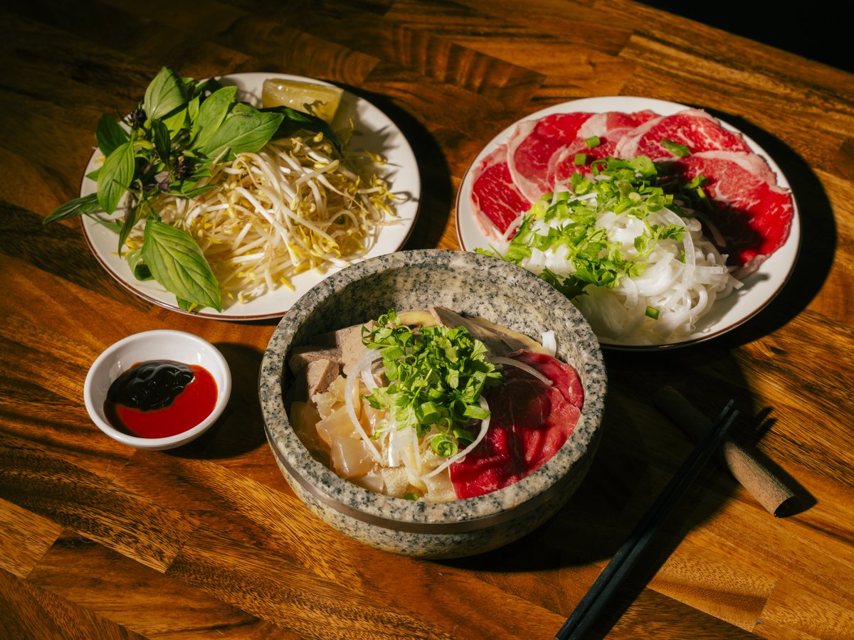 Bà Năm stone-bowl phở spread — rare beef, fresh herbs, bean sprouts and house condiments on a timber table