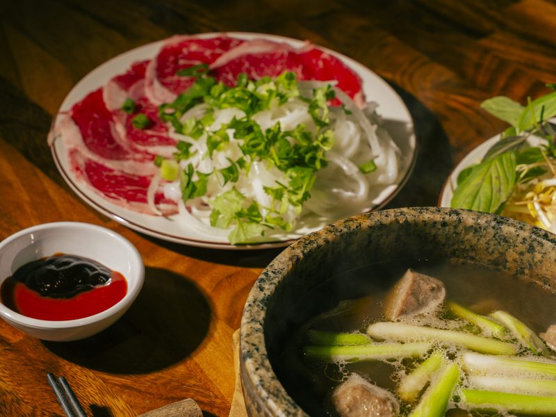 Live phở service — steaming broth over rare beef with a plate of paper-thin slices waiting