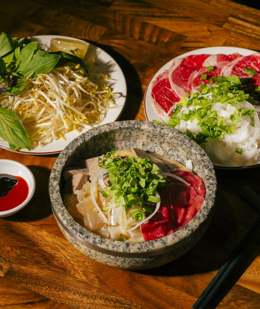 Bà Năm stone-bowl phở spread — rare beef, fresh herbs, bean sprouts and house condiments on a timber table